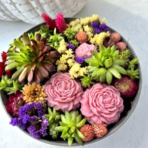 Op de afbeelding: Een decoratief arrangement van vetplanten en gedroogde bloemen in een grijze kom. Het arrangement bevat groene, paarse, gele en roze bloemen, met twee grote, gebeeldhouwde roze rozen als middelpunt. De kom is rond en gevuld met verschillende texturen en kleuren.