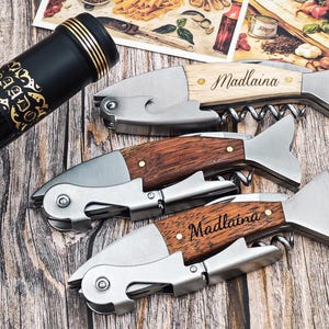 May include: Three fish-shaped corkscrews with wooden handles and stainless steel components are displayed on a wooden surface. The handles are engraved with the name "Madlaina". A black wine bottle with gold accents is also visible.