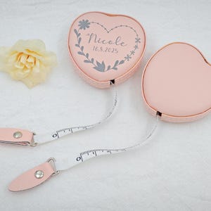 May include: Two pink heart-shaped measuring tapes with white measuring tape extending from each. One tape has a floral design and the name "Nicole" with the date "16.8.2025". A yellow flower is in the background.