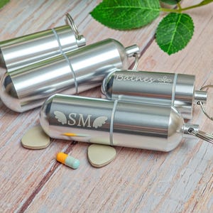 May include: Silver pill fobs with keychains, some engraved with initials and names. One pill fob has the initials "SM" with wing designs. A capsule-shaped pill and two beige shapes are next to one of the pill fobs.