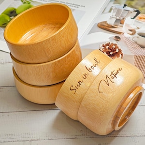 May include: A stack of four wooden bowls with a natural finish. One bowl is tilted, revealing the text "Sun in bowl Nature" etched on its side. The bowls are arranged on a light-colored wooden surface.