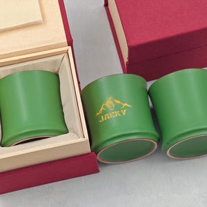 May include: Three green ceramic mugs, one inside a wooden box with a red lid. One mug has a gold mountain logo and the word "JACKY". The mugs have a curved design and a light brown base.