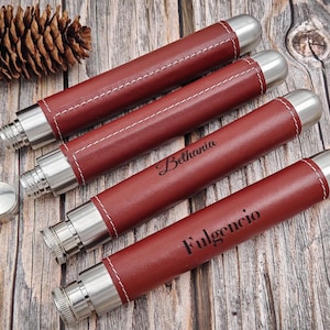 May include: Four cylindrical flasks with claret leather sleeves and stainless steel accents. Each flask is personalised with a name in black script. White stitching details the leather. Two small, silver caps are visible.