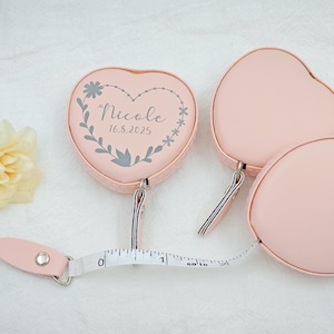 May include: Pink heart-shaped measuring tapes with a floral design and the name "Nicole" and the date "16.8.2025" printed on one. The tape measure extends to 152 cm. The other two tapes are plain pink hearts.