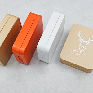 Może przedstawiać: Cztery prostokątne pudełka w różnych kolorach: beżowym, pomarańczowym i białym. Jedno beżowe pudełko ma złote wykończenie. Czwarte pudełko jest beżowe z grafiką głowy jelenia i napisem "Dachin". Pudełka są zamknięte.