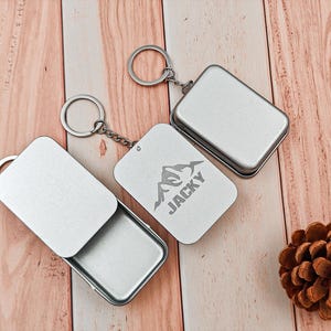 May include: Three silver rectangular keyrings, one engraved with a mountain graphic and the name "JACKY". One keyring is open, showing the inside. They are displayed on a wooden surface.