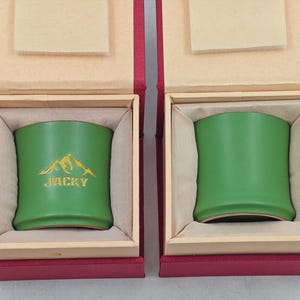 May include: Two green cylindrical containers with a curved design, nestled in red and beige boxes. One container has a gold mountain graphic and the name "JACKY". The boxes are open, revealing the containers inside.