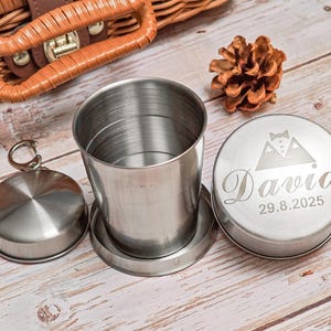 Puede incluir: Una taza plegable de acero inoxidable con una tapa a juego y un pequeño recipiente redondo. La tapa del recipiente está grabada con el nombre "David" y la fecha "29.8.2025". Una piña se encuentra cerca.