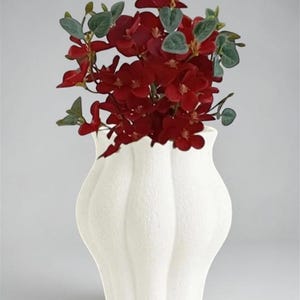 May include: A white ceramic vase with a textured surface, holding vibrant red flowers and green foliage. The vase has a unique, wavy design and is filled with a lush floral arrangement, creating a striking contrast.