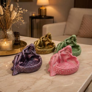 May include: Four decorative trays in purple, gold, green, and pink, each featuring a sculpted figure of a reclining person resting on one side. The trays have a textured, pebble-like interior surface and a smooth, contoured outer edge design.