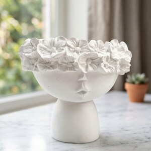 Puede incluir: Una maceta de cer&aacute;mica blanca con forma de rostro y una corona floral. La maceta tiene forma de cuenco redondo con un dise&ntilde;o de rostro y una base de pedestal. La corona floral est&aacute; hecha de flores blancas detalladas. La maceta est&aacute; sobre una superficie de m&aacute;rmol.