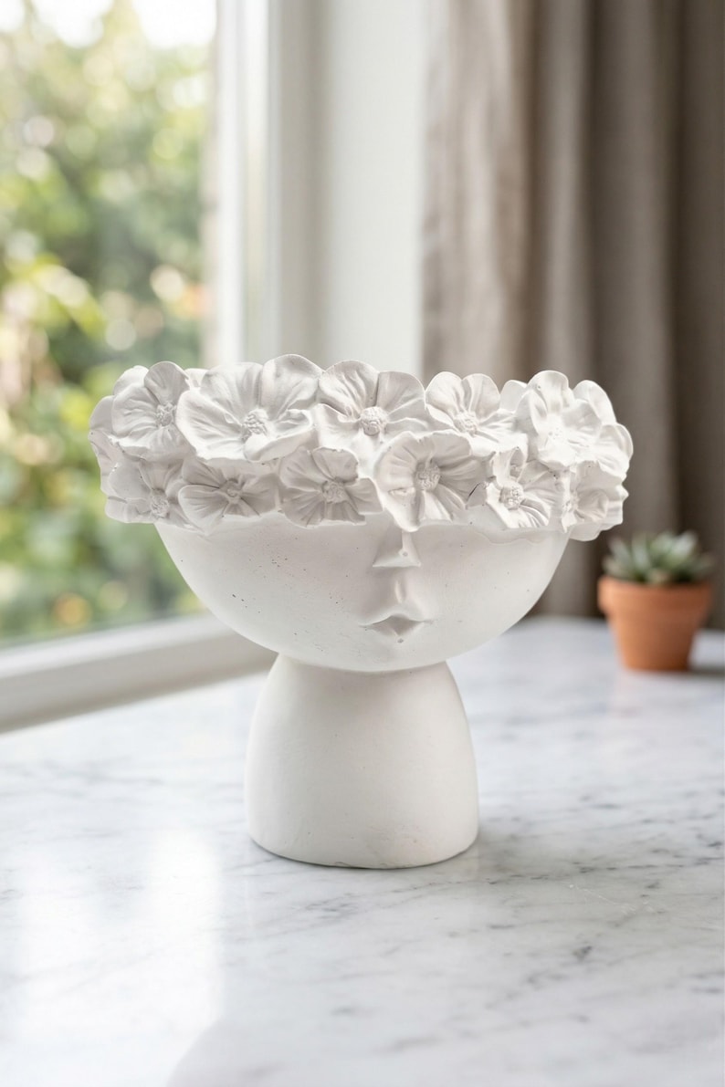 Puede incluir: Una maceta de cer&aacute;mica blanca con forma de rostro y una corona floral. La maceta tiene forma de cuenco redondo con un dise&ntilde;o de rostro y una base de pedestal. La corona floral est&aacute; hecha de flores blancas detalladas. La maceta est&aacute; sobre una superficie de m&aacute;rmol.