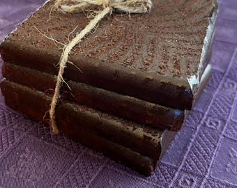 Può includere: Una pila di quattro sottobicchieri quadrati marrone scuro, legati con spago. I sottobicchieri hanno una superficie strutturata e sono impilati su una tovaglia viola a fantasia. Lo spago è legato a fiocco.