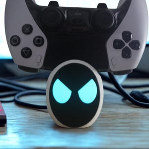 Può includere: Un controller di gioco bianco e nero è posizionato sopra un piccolo dispositivo ovale con occhi blu luminosi. Il controller ha pulsanti neri e un pad direzionale. Il dispositivo è su una superficie di legno con altri componenti elettronici.