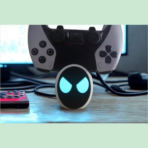 Op de afbeelding: Een klein, ovaalvormig robotapparaat met een zwart gezicht en oplichtende lichtblauwe ogen, geplaatst voor een wit-zwarte videogamecontroller. Het apparaat heeft een gladde witte buitenkant en een donker, rond scherm.