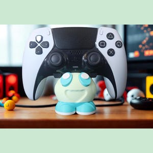 May include: A white and black video game controller rests on a small, light green and blue character-shaped stand. The stand features two large white circular eyes and a wide, curved mouth, providing a stable base for the gaming peripheral.