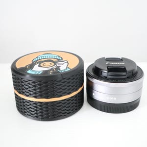 May include: A black and gold container with a cartoon image on the lid, next to a Canon camera lens with a black lens cap. The container has a textured, woven design. The lens is silver and black.