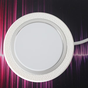 May include: A white wireless charger with a silver ring and a textured outer edge. The charger has a white cable attached. The background has a pattern of red, purple, and black.