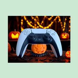 May include: A white and black wireless video game controller resting on top of a small orange jack-o'-lantern figure. The controller features standard directional buttons, two analog sticks, and a central touch pad, positioned against a dark background.