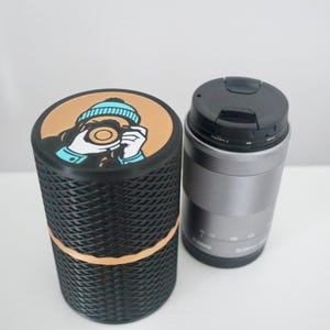 Könnte beinhalten: Ein schwarzer und hellbrauner zylindrischer Behälter mit einer Grafik einer Person, die ein Foto macht, neben einem silbernen Kameraobjektiv mit schwarzer Kappe. Der Behälter hat eine gewebte Textur.