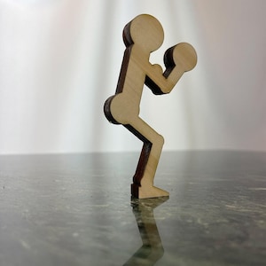 以下が含まれることがあります： 片方の腕を上げて拳を握ったボクシングのポーズをとる人の木製の切り抜き。切り抜きは薄い茶色の木でできており、濃い茶色のアクセントが付けられている。