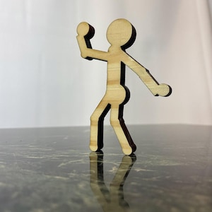 May include: A wooden stick figure figurine in a throwing pose, with a ball in its hand. The figurine is made of light brown wood with dark brown outlines. The figurine is standing on a dark green surface.