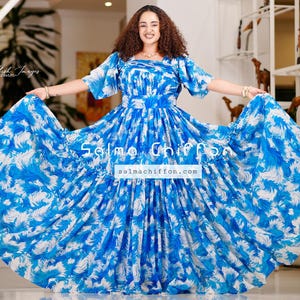 Puede incluir: Una mujer vistiendo un vestido estampado azul y blanco con una falda con volantes. El vestido tiene un escote cuadrado y mangas cortas. El vestido se llama "Salma Chiffon" y el sitio web es salmachiffon.com.