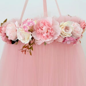 Puede incluir: Un m&oacute;vil colgante con una disposici&oacute;n circular de flores artificiales rosas y crema, incluyendo peon&iacute;as y rosas, acentuadas con peque&ntilde;as hojas verdes. Una tela de tul larga, transparente y de color rosa claro cuelga hacia abajo.