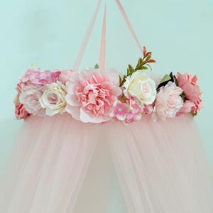 Puede incluir: Un dosel colgante con un marco circular adornado con un arreglo de flores artificiales en tonos rosa, crema y blanco. El dosel est&aacute; hecho de tela transparente de color rosa claro que cae hacia abajo desde el anillo cubierto de flores.