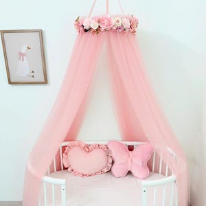 Puede incluir: Un dosel de cama de tul rosa con una corona de flores artificiales de rosas y peon&iacute;as en la parte superior. El dosel cuelga sobre una cuna ovalada blanca que contiene un coj&iacute;n en forma de coraz&oacute;n y otro en forma de mariposa, ambos en rosa.