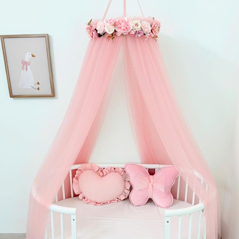 Puede incluir: Un dosel de cama de tul rosa con una corona de flores artificiales de rosas y peon&iacute;as en la parte superior. El dosel cuelga sobre una cuna ovalada blanca que contiene un coj&iacute;n en forma de coraz&oacute;n y otro en forma de mariposa, ambos en rosa.