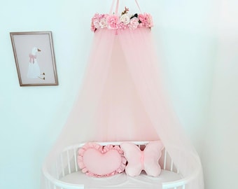 May include: A pale pink tulle bed canopy featuring a crown of artificial pink roses and greenery at the top. Inside the white oval crib, there is a heart-shaped pillow and a butterfly-shaped pillow, both in a matching soft pink fabric.