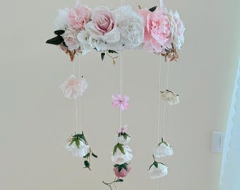 May include: A floral mobile featuring a ring of pink and white flowers, with hanging strands of smaller blossoms and a pink bow. The mobile is suspended by white ribbons, creating a delicate and airy aesthetic.