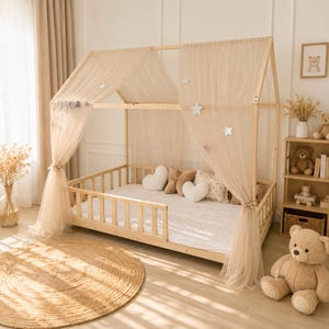 May include: A light-colored wooden house-shaped floor bed frame with a white mattress, surrounded by sheer beige tulle curtains draped over the roof structure. The frame is decorated with white star cutouts and the name MAYA in block letters.