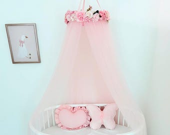 May include: A pale pink bed canopy featuring a crown of artificial flowers in shades of pink and white. Inside the white oval crib, there is a heart-shaped pillow and a butterfly-shaped pillow, both in a matching soft pink fabric.