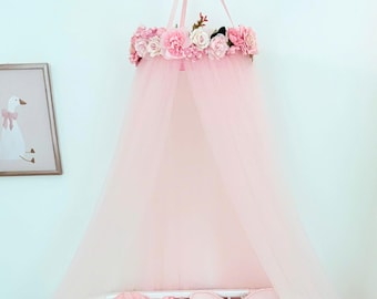 May include: A pale pink tulle bed canopy featuring a circular top adorned with a ring of artificial pink and white roses and peonies. The sheer fabric hangs from a central suspension point, creating a draped effect over a white crib frame.
