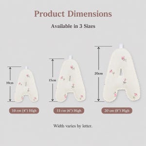 May include: Three off-white fabric letter A ornaments with a small pink floral pattern, featuring scalloped edges and a hanging loop. The image displays three sizes: 10 cm (4 inches), 15 cm (6 inches), and 20 cm (8 inches) in height.