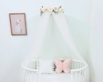May include: A white oval crib with a sheer white canopy topped with a floral arrangement of pink and white roses. Inside the crib are a square white ruffled pillow and a light pink butterfly-shaped pillow. A framed print of a goose hangs nearby.