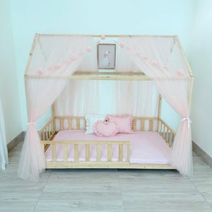 May include: A light wood-framed bed with a canopy of sheer pink fabric. The bed is decorated with pink pillows and a heart-shaped pillow. A framed picture hangs above the pillows, and pink pom-poms adorn the canopy.