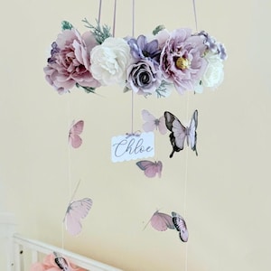 May include: A floral baby mobile featuring a ring of purple, pink, and white flowers. Butterflies in shades of pink and black dangle below, with a tag that reads "Chloe". The mobile hangs from purple ribbons, creating a whimsical nursery decoration.