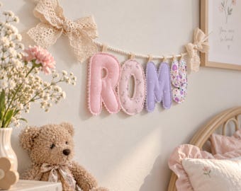 May include: A decorative fabric banner spelling the name ROMY in soft pastel colors, featuring individual stuffed letters suspended from a thick white rope adorned with two large, intricate cream-colored lace bows at each end.