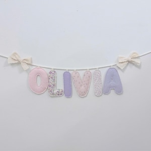May include: A fabric letter banner spelling the name OLIVIA, featuring individual stuffed letters in shades of pink, purple, and floral patterns, suspended on a white cord with a cream-colored fabric bow tied at each end of the string.