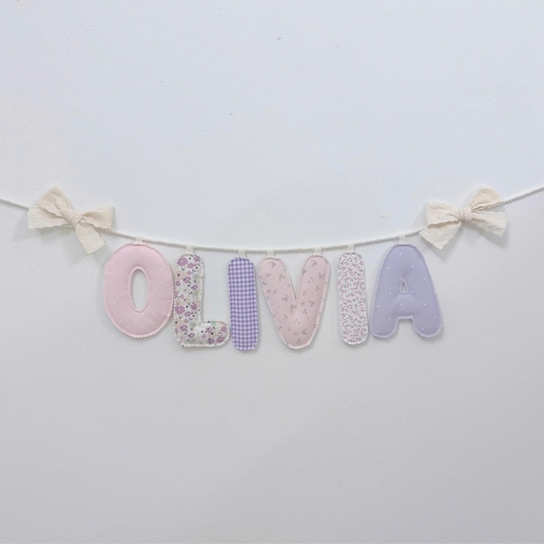May include: A fabric letter banner spelling the name OLIVIA, featuring individual stuffed letters in shades of pink, purple, and floral patterns, suspended on a white cord with a cream-colored fabric bow tied at each end of the string.