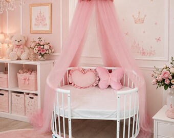 May include: A white oval crib with a pink tulle canopy featuring a floral crown. Inside the crib are a heart-shaped pillow and a butterfly-shaped pillow, both in light pink. The crib has a white frame with wheels and a smooth white mattress.