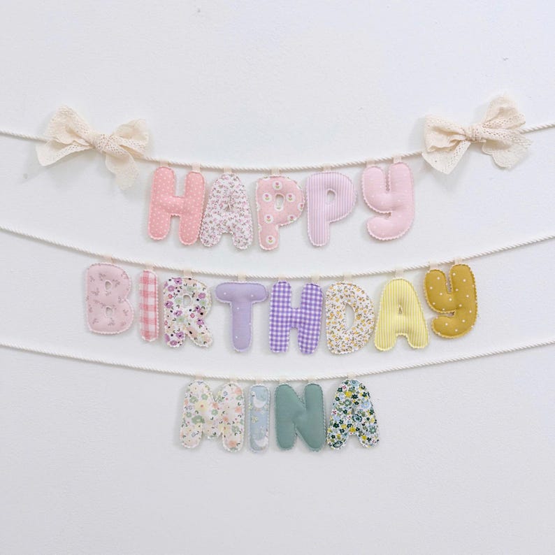 May include: A fabric banner featuring three rows of stuffed, padded letters hanging from white ropes. The letters spell out HAPPY BIRTHDAY MINA in various pastel colors, floral patterns, gingham, and polka dots, accented by two cream lace bows.