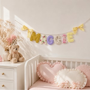 May include: A fabric letter garland spelling MAGGIE, featuring individual stuffed letters in mustard yellow, floral, purple gingham, and cream patterns, suspended on a string with yellow ribbon bows at each end.