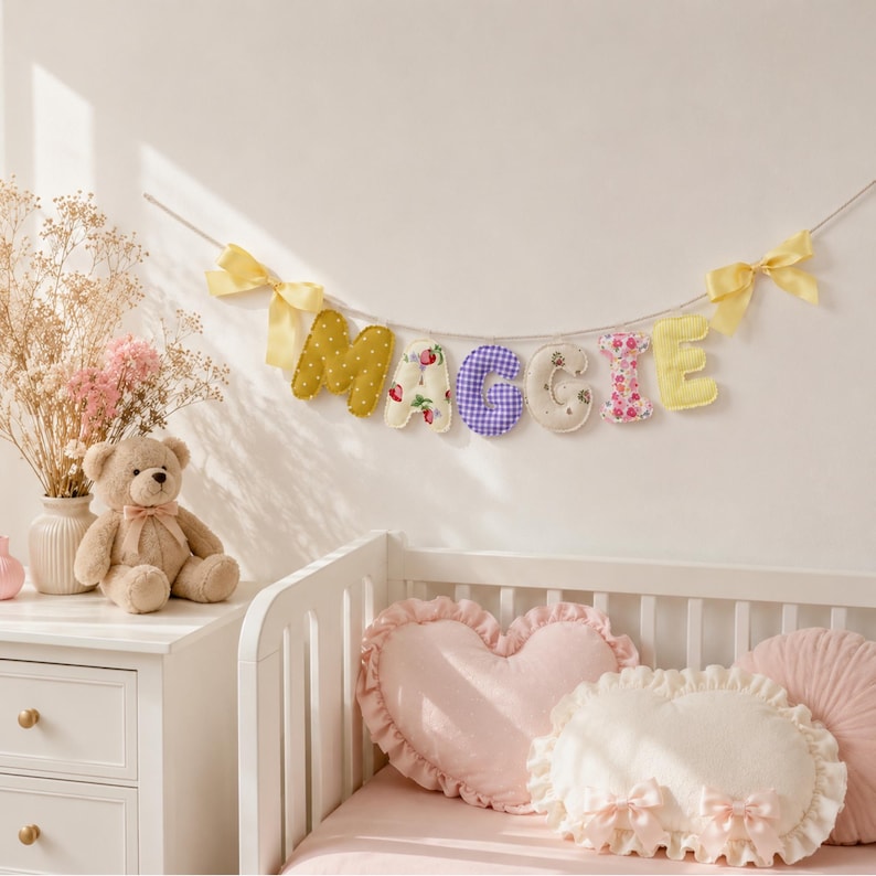 May include: A fabric letter garland spelling MAGGIE, featuring individual stuffed letters in mustard yellow, floral, purple gingham, and cream patterns, suspended on a string with yellow ribbon bows at each end.