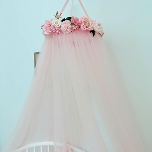 Puede incluir: Un dosel de tul rosa p&aacute;lido con una parte superior circular adornada con un grupo de flores artificiales en tonos rosa claro, crema y blanco. La tela transparente cae desde la corona floral, creando una apariencia suave y superpuesta.