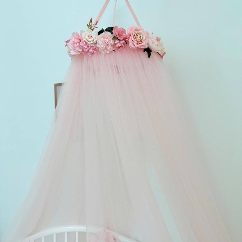 Puede incluir: Un dosel de tul rosa p&aacute;lido con una parte superior circular adornada con un grupo de flores artificiales en tonos rosa claro, crema y blanco. La tela transparente cae desde la corona floral, creando una apariencia suave y superpuesta.