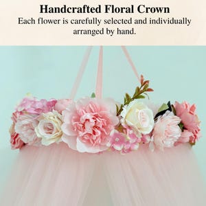 Puede incluir: Una corona floral con flores artificiales rosas y crema dispuestas en una banda circular, con largas cintas rosas para colgar. Debajo de las flores, cuelga un dosel de tul rosa transparente. El texto dice: Handcrafted Floral Crown.